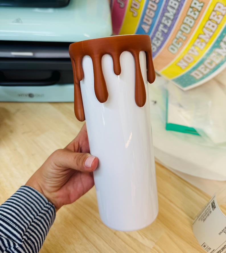May include: A white cylindrical tumbler featuring a brown drip design around the top rim. The brown material appears to be a textured, raised coating that mimics liquid dripping down the sides of the smooth, white surface of the drink container.
