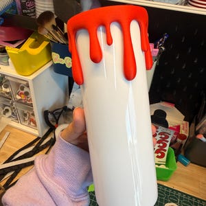 May include: A white cylindrical tumbler featuring a textured, bright orange drip design around the top rim. The surface of the tumbler is smooth and glossy, while the orange drips have a matte, raised appearance that mimics liquid flowing downward.