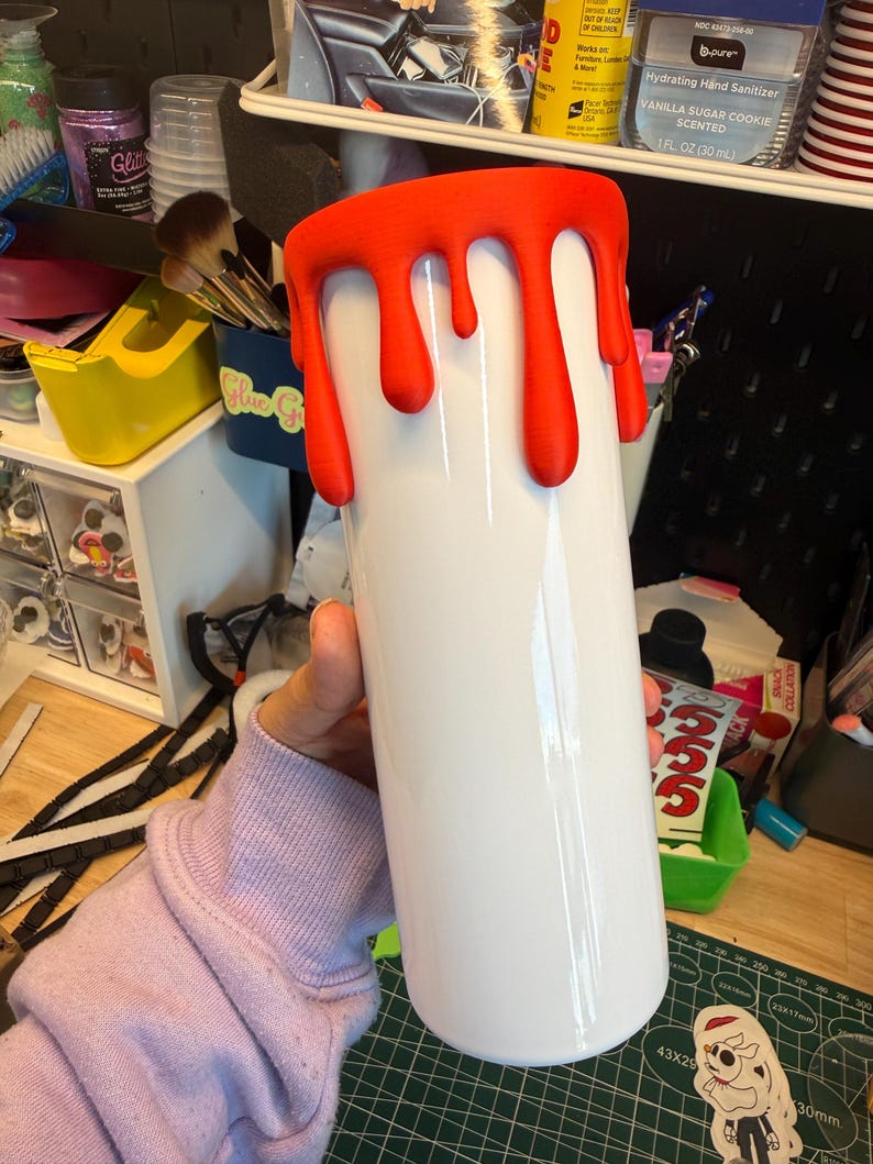 May include: A white cylindrical tumbler featuring a textured, bright orange drip design around the top rim. The surface of the tumbler is smooth and glossy, while the orange drips have a matte, raised appearance that mimics liquid flowing downward.