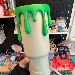 May include: A beige tumbler featuring a bright green slime drip effect around the top rim. The cup has a cylindrical body that tapers toward the base, with a smooth matte finish on the beige surface and a raised texture on the green slime.