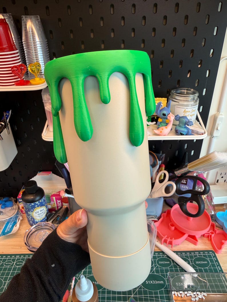 May include: A beige tumbler featuring a bright green slime drip effect around the top rim. The cup has a cylindrical body that tapers toward the base, with a smooth matte finish on the beige surface and a raised texture on the green slime.