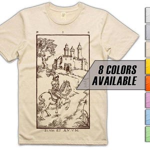 Può includere: T-shirt beige con un'illustrazione in bianco e nero di un cavaliere a cavallo davanti a un castello. La grafica include testo e la frase "8 COLORS AVAILABLE" in un banner grigio. La maglietta è disponibile in vari colori.
