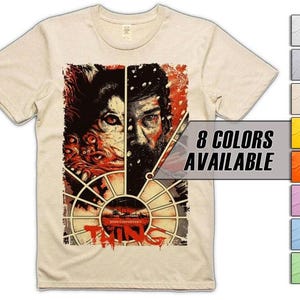 May include: A cream-colored t-shirt featuring a movie poster design. The graphic is split vertically, showing a creature on the left and a man on the right. The text "THE THING" is at the bottom. The text "8 COLORS AVAILABLE" is also present.