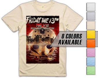 Puede incluir: Camiseta beige con un gráfico de la película "Viernes 13 Parte VIII: Jason Takes Manhattan". El diseño incluye el título de la película, un paisaje urbano y la máscara de hockey de Jason. También se muestra el texto "8 COLORES DISPONIBLES".