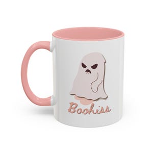 May include: White ceramic mug with a pink interior and handle. The mug features a cartoon ghost with an angry expression and the word "Boohiss" in a cursive font. A fun and spooky design.