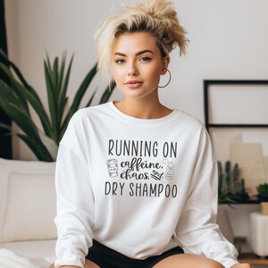 May include: White sweatshirt with the text "RUNNING ON caffeine, chaos, and DRY SHAMPOO" in black. The text is accompanied by a drawing of a coffee cup and a can of dry shampoo.