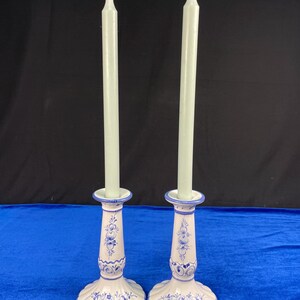 May include: Two white ceramic candle holders with blue floral designs, each holding a tall, pale green candle. The candle holders have a flared base and a detailed, hand-painted appearance, perfect for home decor.