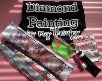 Może przedstawiać: Czarna koszulka z białym napisem Diamond Painting is my therapy, w zestawie z kompletem do malowania diamentami zawierającym zadrukowane płótno, zieloną tackę, różowy aplikator, kwadrat wosku oraz saszetkę gorącej czekolady Nestle.