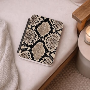 May include: A rectangular electronic tablet case featuring a high-contrast snakeskin pattern in shades of beige, tan, and black. The cover has a smooth, flat surface with a black spine edge and rounded corners, displaying a repeating scale texture.