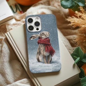 May include: A blue smartphone case featuring a detailed illustration of a brown hare wearing a red scarf, standing in a snowy landscape with falling snowflakes. The case has a cutout for a triple-lens camera system on the upper left corner.