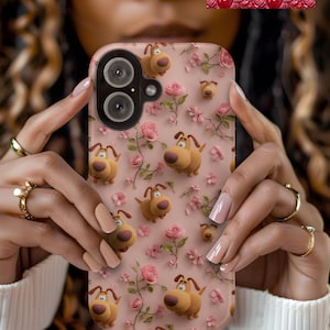 May include: A pink phone case with a repeating pattern of cartoon dogs and pink roses. The case has a camera cutout. The design is whimsical and floral. The case is held by a person.