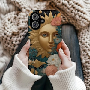 May include: A phone case featuring a golden sun face with blue eyes, surrounded by a floral arrangement of pink, orange, and white flowers. The case has a black background with a starry design.