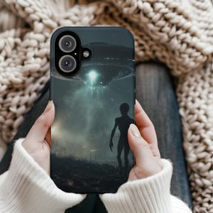 May include: A black phone case featuring a graphic of a UFO and a shadowy figure standing in front of it. The case has a camera lens cutout.