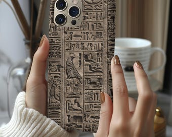 May include: A phone case with a detailed, ancient Egyptian hieroglyphic design in shades of brown and tan. The case has a textured appearance and features a camera module. The phone is held by a person wearing a white sweater.