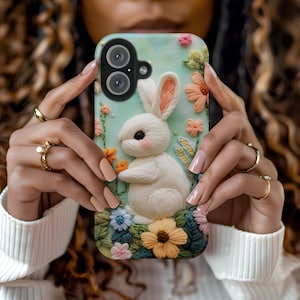 May include: A white bunny embroidered on a phone case with a floral design. The phone case has a light blue background with pink, yellow, and green flowers. The bunny is holding a small pink flower.