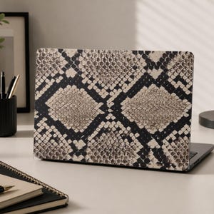 May include: A laptop computer featuring a protective skin with a high-contrast snakeskin pattern. The design consists of repeating diamond shapes in shades of beige, tan, and dark brown, set against a deep black background, covering the entire lid.