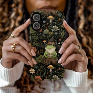 May include: A black phone case featuring a whimsical illustration of a green frog wearing a witch hat, surrounded by mushrooms and floral elements. The case has a dark background with a detailed botanical pattern.