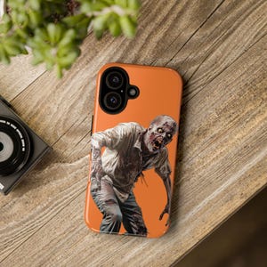 May include: Orange phone case featuring an illustration of a zombie. The zombie is wearing a white shirt and has a bloody face.