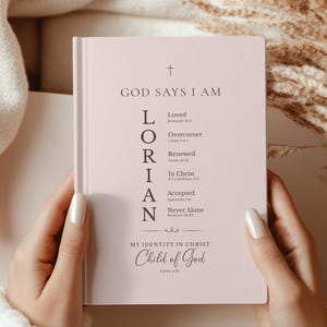 May include: A light pink hardcover journal featuring the text GOD SAYS I AM, a vertical acrostic for the name LORIAN with biblical references, and the phrase MY IDENTITY IN CHRIST Child of God printed in dark gray ink on the front cover.