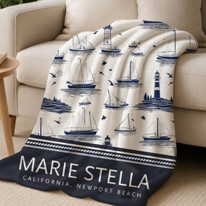 May include: A cream-coloured throw blanket with a nautical design of navy blue sailboats, lighthouses, and birds. The blanket has a navy blue border with the text "MARIE STELLA CALIFORNIA, NEWPORT BEACH."
