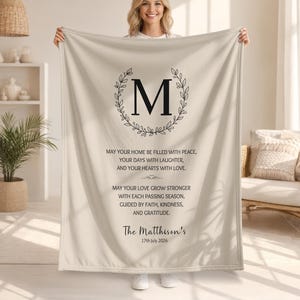 May include: A beige fleece blanket featuring a large black letter M centred within a leafy wreath. Below the letter, black text reads: May your home be filled with peace, your days with laughter, and your hearts with love. The Matthison's 17th July 2026.