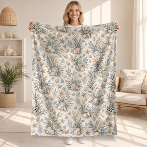 May include: A rectangular throw blanket featuring a repeating pattern of seashells, starfish, and blue and tan botanical sprigs on a cream-coloured background. The design includes various types of shells and sand dollars arranged in clusters.