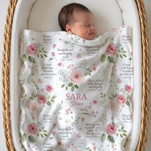 May include: A white baby blanket featuring a repeating pattern of pink roses, green leaves, and various religious scripture verses. The name Sara Grace is printed in the center in pink text. The blanket is draped over a sleeping infant.