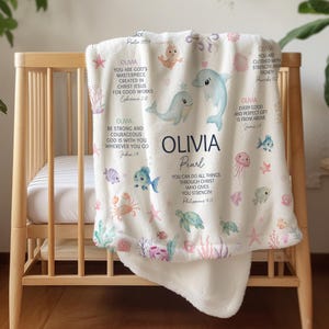 Puede incluir: Una manta de forro polar blanca con el nombre personalizado Olivia Pearl, versículos bíblicos e ilustraciones coloridas de criaturas marinas que incluyen un delfín, ballena, tortuga, cangrejo y medusa, dispuestas en un patrón.