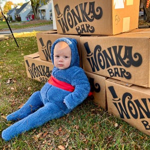 May include: A baby dressed as Willy Wonka from the movie Willy Wonka and the Chocolate Factory. The baby is wearing a blue fuzzy jumpsuit with a red sash. The baby is sitting in front of a stack of cardboard boxes with the words "Wonka Bar" printed on them.