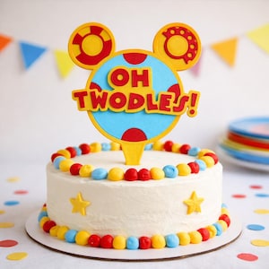 May include: A round, white frosted cake decorated with red, yellow, and blue sweets. The cake is topped with a yellow and red cake topper that reads "OH TWODLES!". Yellow star decorations are also on the cake, perfect for a birthday.