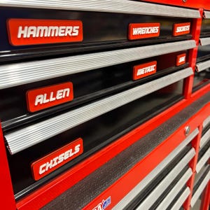 May include: A red and black tool chest with labeled drawers. The labels, in white, read "HAMMERS", "WRENCHES", "SOCKETS", "ALLEN", "METRIC", and "CHISELS". The drawers have silver trim.