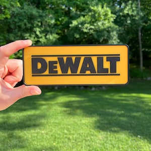 Puede incluir: Un letrero rectangular amarillo y negro con la palabra "DEWALT" en letras negras en negrita. El letrero se sostiene contra un fondo de hierba verde y árboles.