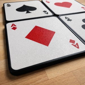 May include: Four square coasters with playing card designs. The coasters feature the Ace of Spades, Ace of Hearts, Ace of Clubs, and Ace of Diamonds. Each coaster has a white background with a black border.