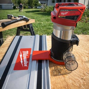 Puede incluir: Una fresadora compacta Milwaukee roja y plateada con un mango negro y un puerto transparente de recogida de polvo. La fresadora está montada sobre un riel plateado con una guía roja. Dos martillos descansan sobre una superficie de madera.