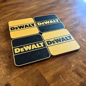 Puede incluir: Cuatro posavasos cuadrados con esquinas redondeadas. Cada posavasos tiene un fondo negro con la palabra "DEWALT" en letras amarillas en negrita. Los posavasos son amarillos con letras negras y los bordes son amarillos.