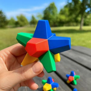 May include: A colorful star-shaped puzzle toy with red, blue, green, and yellow geometric shapes. The toy is held in a hand, with additional similar toys visible in the background. The background features a green lawn and trees.