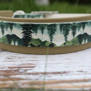 K&ouml;nnte beinhalten: Ein braunes Hundehalsband mit einem wei&szlig;en und gr&uuml;nen Berg- und Waldmuster. Das Halsband ist aus strapazierf&auml;higem Material gefertigt und hat eine robuste Schnalle.