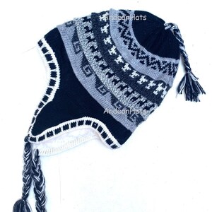 Könnte beinhalten: Eine schwarz-grau-weiße Strickmütze mit Ohrenklappen und geflochtenen Quasten. Die Mütze hat ein geometrisches Muster und den Text "Andean Hats". Die Ohrenklappen sind mit weißem Stoff gefüttert.