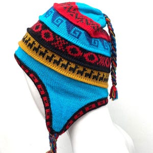 Puede incluir: Un gorro de punto colorido con orejeras y una borla trenzada. El gorro presenta bandas turquesas, rojas, negras y amarillas con patrones geométricos. El gorro está diseñado para mantener la cabeza y las orejas calientes.