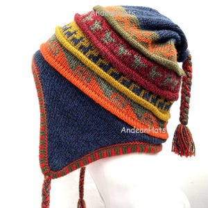 Puede incluir: Un gorro de punto colorido con orejeras y borlas trenzadas. El gorro presenta un patrón de diseños geométricos naranjas, rojos, amarillos y verdes sobre un fondo azul. El texto "Andean Hats" es visible.
