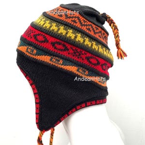 Puede incluir: Un gorro negro tejido con orejeras y una borla trenzada. El gorro presenta un colorido patrón geométrico en rojo, amarillo, naranja y verde. El texto "Andean Hats" es visible en el gorro.