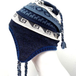 Può includere: Un cappello lavorato a maglia blu, bianco e grigio con paraorecchie e lacci intrecciati. Il cappello presenta motivi geometrici e il testo "Andean Hats". Il cappello è esposto su una testa di manichino bianca.