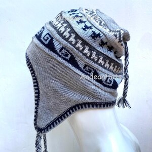 May include: A grey and navy blue knitted hat with ear flaps and braided tassels. The hat features geometric patterns and designs, including stylized animal figures. The hat is made of a soft, textured material, and the ear flaps are trimmed with a dark blue border.