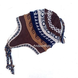 May include: A brown, blue, and cream-coloured knit hat with ear flaps and a braided tassel. The hat features geometric patterns and the text "AndeanHats". This hat is designed for cold weather and is likely handmade.