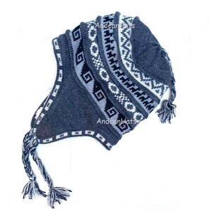 Può includere: Un cappello lavorato a maglia grigio e bianco con paraorecchie e nappe. Il cappello presenta motivi geometrici bianchi, neri e grigi. Il testo "Andean Hats" è visibile sul cappello.