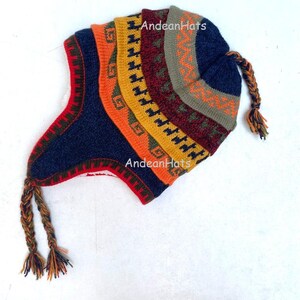 May include: A knitted hat with ear flaps and braided tassels, featuring a vibrant pattern. The hat displays sections of orange, yellow, red, and blue with geometric patterns. The text "Andean Hats" is visible on the hat.