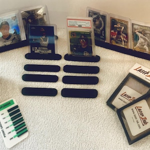 Puede incluir: Un conjunto de accesorios para exhibir tarjetas deportivas, que incluyen soportes de exhibición negros, porta tarjetas y un medidor de puntos. Las tarjetas están en estuches protectores, y algunas están clasificadas. El logotipo "Laser Ink" es visible.