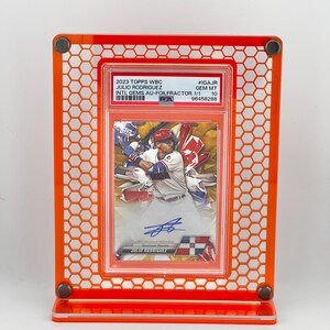 Puede incluir: Un expositor acr&iacute;lico naranja que contiene una tarjeta de b&eacute;isbol. La tarjeta presenta a Julio Rodr&iacute;guez y el texto "2023 TOPPS WBC". El expositor tiene un patr&oacute;n de panal y est&aacute; dise&ntilde;ado para exhibir la tarjeta.