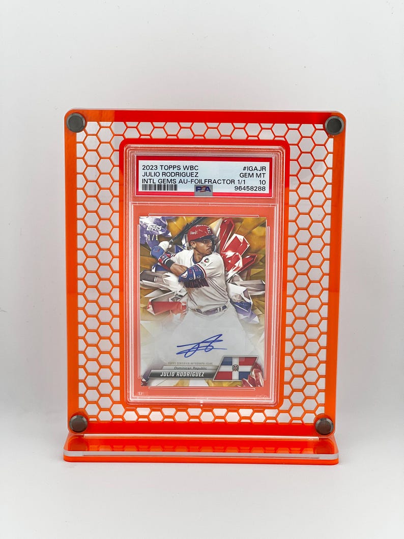 Puede incluir: Un expositor acr&iacute;lico naranja que contiene una tarjeta de b&eacute;isbol. La tarjeta presenta a Julio Rodr&iacute;guez y el texto "2023 TOPPS WBC". El expositor tiene un patr&oacute;n de panal y est&aacute; dise&ntilde;ado para exhibir la tarjeta.
