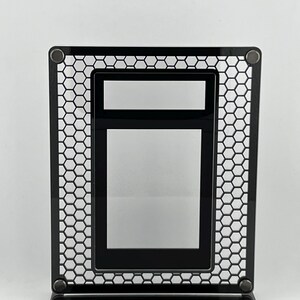 Puede incluir: Un marco de fotos de metal negro con un dise&ntilde;o de malla de panal. El marco tiene forma rectangular con una abertura rectangular m&aacute;s peque&ntilde;a en el interior. El marco est&aacute; soportado por una base con una franja blanca.